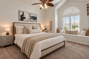 A bedroom with a large bed, a ceiling fan, two bedside tables with lamps, a window seat with pillows, and artwork on the wall.