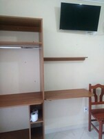 Una habitación con un televisor montado en la pared, estantes de madera, una silla y un rollo de papel higiénico sobre uno de los estantes.