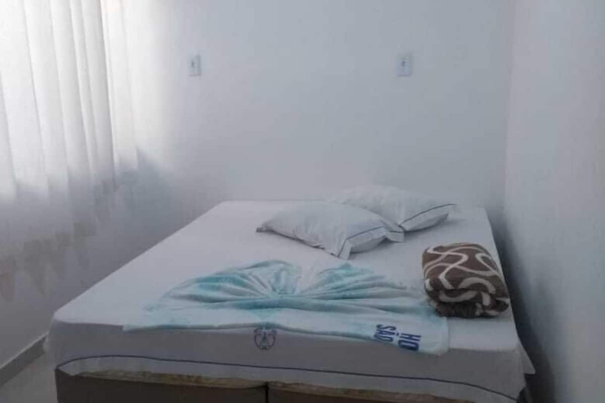 Una habitación pequeña con una cama individual, pared blanca lisa, ventana con persianas y un colchón con manta azul.