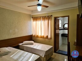 Habitación de hotel con dos camas individuales, ventilador de techo, ventana con cortinas y vistas al baño y la cocina a través de una puerta abierta.