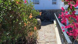 Uma trilha de pedra leva a um edifício branco com uma janela, cercado por vegetação exuberante e flores cor-de-rosa.