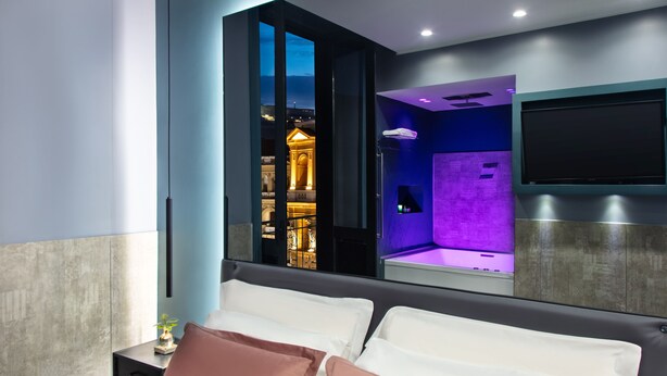 A modern hotel room with a large bed, a TV mounted on the wall, and a view of a lit-up building outside. - Enjoy Art (Naples)