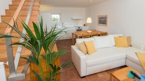 A modern living room with a white sofa, a dining table, and a staircase leading to a kitchen. - Convento das Bernardas Lux T2 Triplex (Tavira)