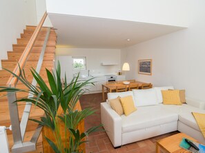 A modern living room with a white sofa, a dining table, and a staircase leading to a kitchen. - Convento das Bernardas Lux T2 Triplex (Tavira)