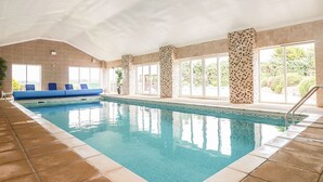 Una piscina interior con agua azul cristalina, un área de descanso azul y amplios ventanales con vistas al exterior.
