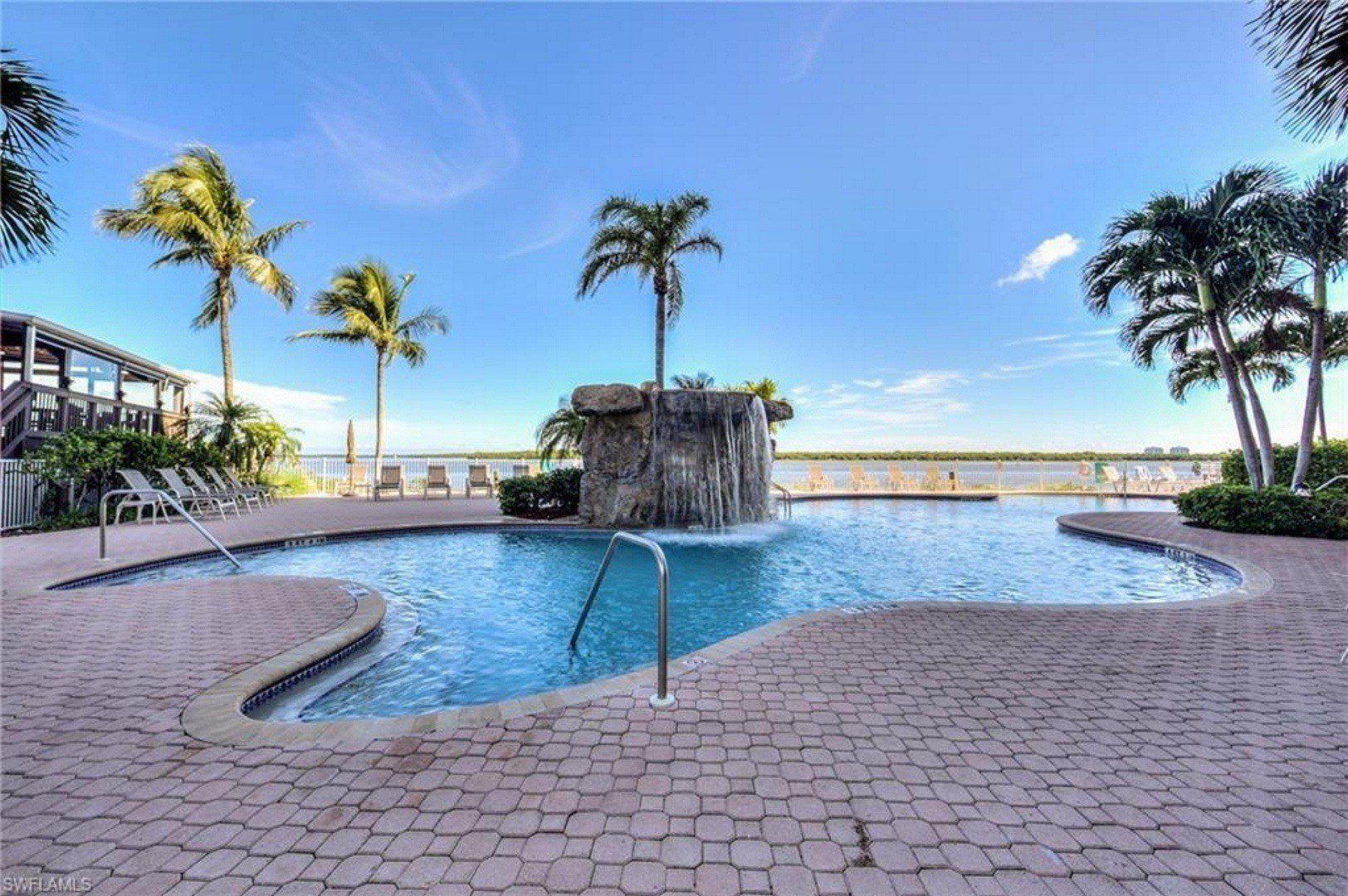 A swimming pool with a waterfall feature, surrounded by palm trees and lounge chairs.