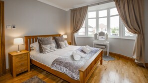 A neatly made bed with a grey bedspread and white pillows, a wooden nightstand with a lamp, and a window with curtains. - Family House (Weymouth)