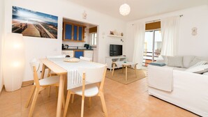 Een woonkamer met een bank, een eettafel met stoelen, een keuken met kasten, een televisie en een groot raam met gordijnen.