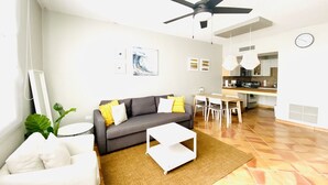 A modern living room with a gray sofa, white armchair, and a coffee table. A ceiling fan and a dining area with a kitchen are visible in the background. - Oceanview Villa - Steps Away from the Beach / Palmas del Mar Resort. (Palmas del Mar)