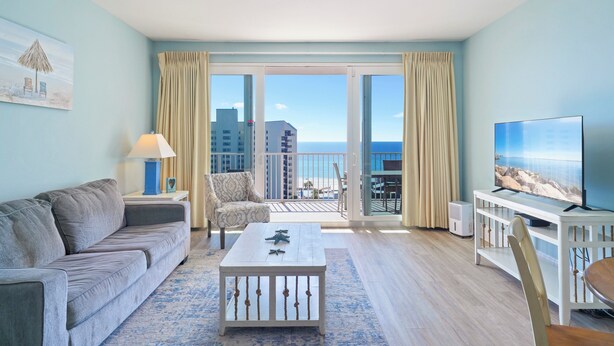 A living room with a sofa, armchair, coffee table, TV, and balcony with curtains.