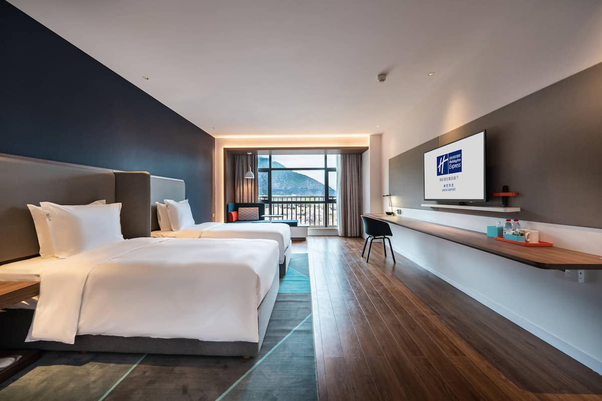 A hotel room with a large bed, a desk, a TV, and a balcony with a mountain view.