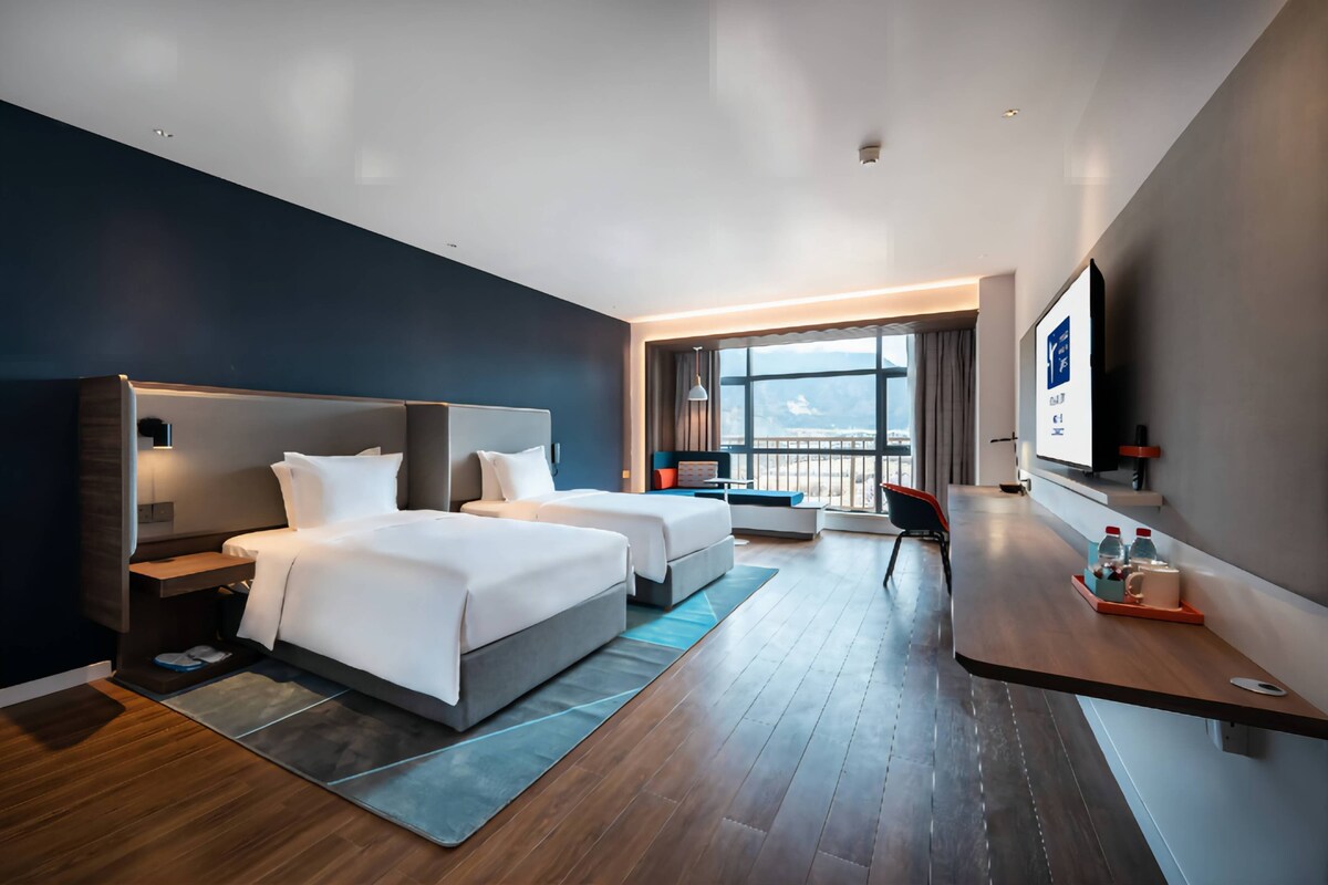 A modern hotel room with two beds, a desk, a TV, and a balcony with a view.