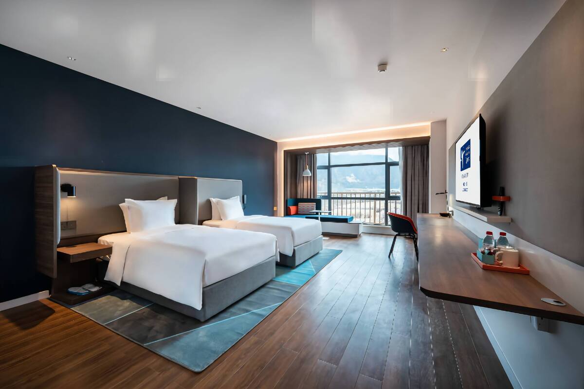 A modern hotel room with two beds, a desk, a TV, and a balcony with a view.