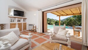 A spacious living room with a large TV, wooden furniture, and a view of a pool and mountains. - Casa Chenal 10 Emma Villas (Arzachena)