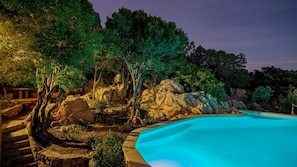 Uma piscina rodeada por rochas e árvores, com um deck de madeira e degraus de pedra que dão acesso à piscina.