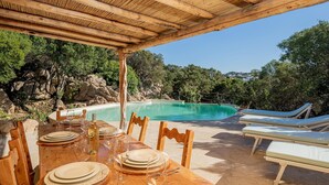A poolside dining area with a wooden table set for a meal, surrounded by lounge chairs and a view of a clear blue pool. - Casa Chenal 10 Emma Villas (Arzachena)