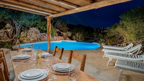 Uma área de refeições ao ar livre com piscina, pérgola de madeira e espreguiçadeiras.