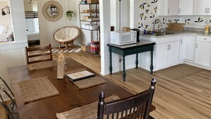 Ein Essbereich mit Holztisch, Stühlen und einer Chandelier. Es gibt eine Küche mit weißen Schränken und einer Mikrowelle auf einem kleinen Tisch.