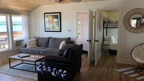 Ein geräumiges Wohnzimmer mit grauem Sofa, einem schwarzen Hocker und einem Couchtisch. Es gibt einen Deckenventilator, ein gerahmtes Gemälde und einen Blick auf einen Balkon mit einer Holzterrasse.
