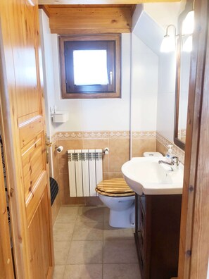 A compact bathroom with a wooden door, a toilet with a wooden seat, a white sink, a mirror, and a window above the toilet.