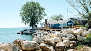A coastal house with a wooden deck, large rocks in the foreground, and a clear view of the sea.