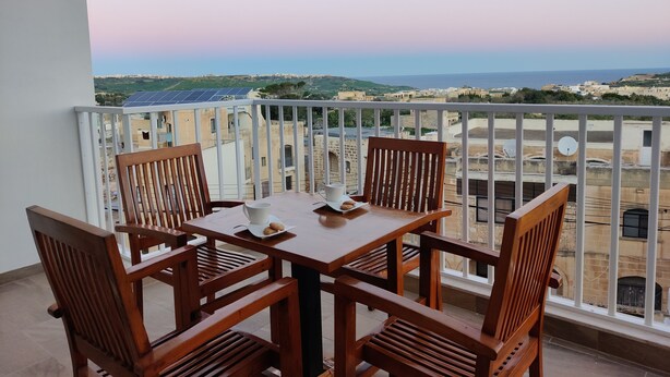 A balcony with a table and chairs, offering a view of the sea and buildings. - Mediterranean Breeze Apartment (Xaghra)