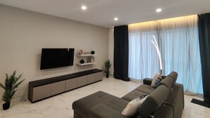 A modern living room with a flat-screen TV mounted on the wall, a sofa, a console table, and a large window with curtains. - Mediterranean Breeze Apartment (Xaghra)