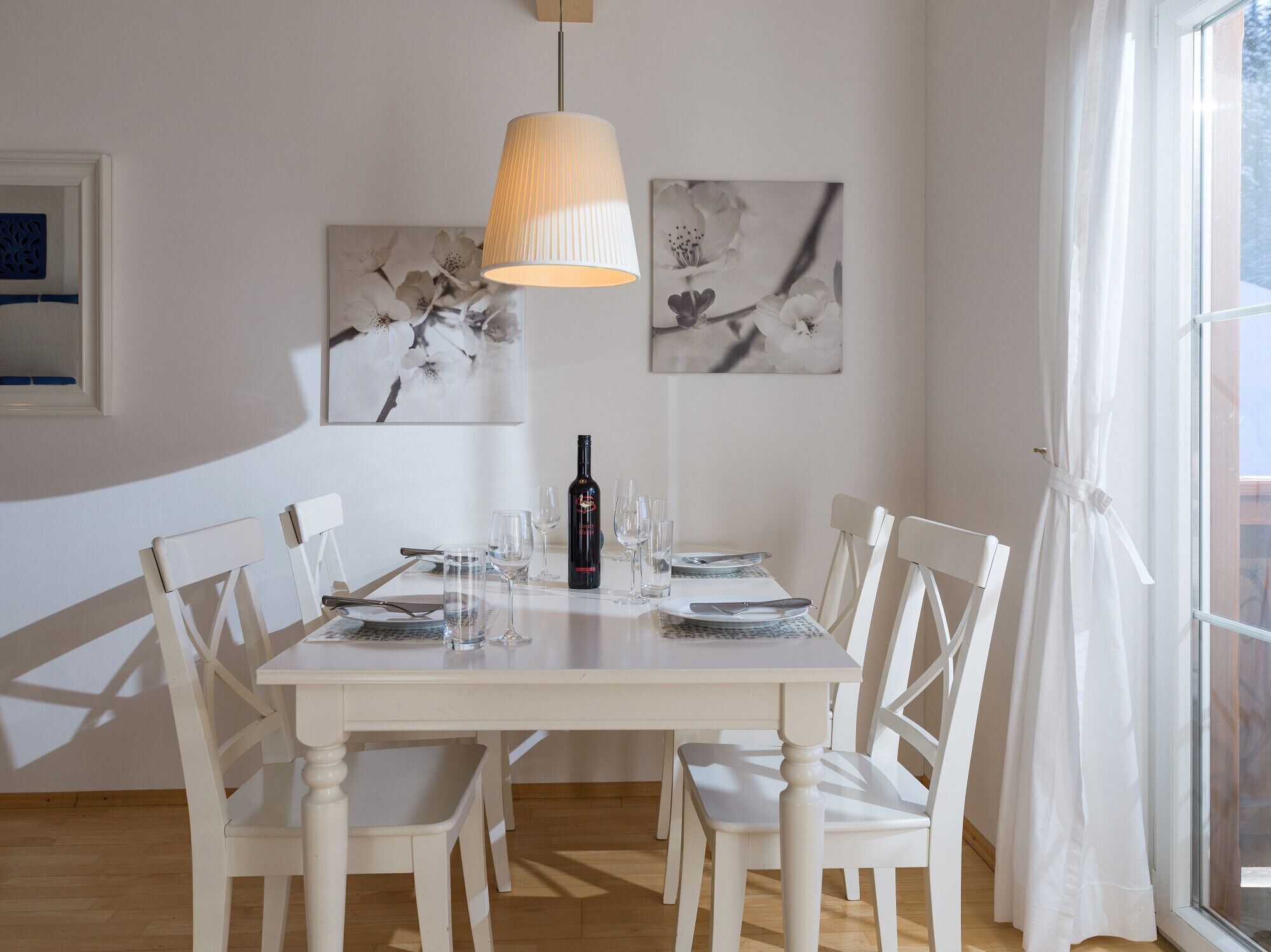 Et spisebord med hvide stole, en vinflaske og glas. To billeder på væggen viser blomster.