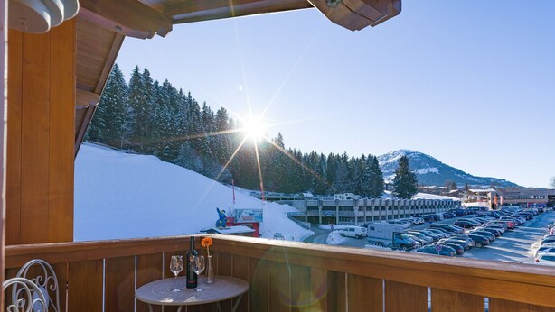A wooden balcony with a table and chairs, overlooking a snowy landscape with a parking area and buildings in the background. - Ski-in / Ski-out (Kirchberg in Tirol)