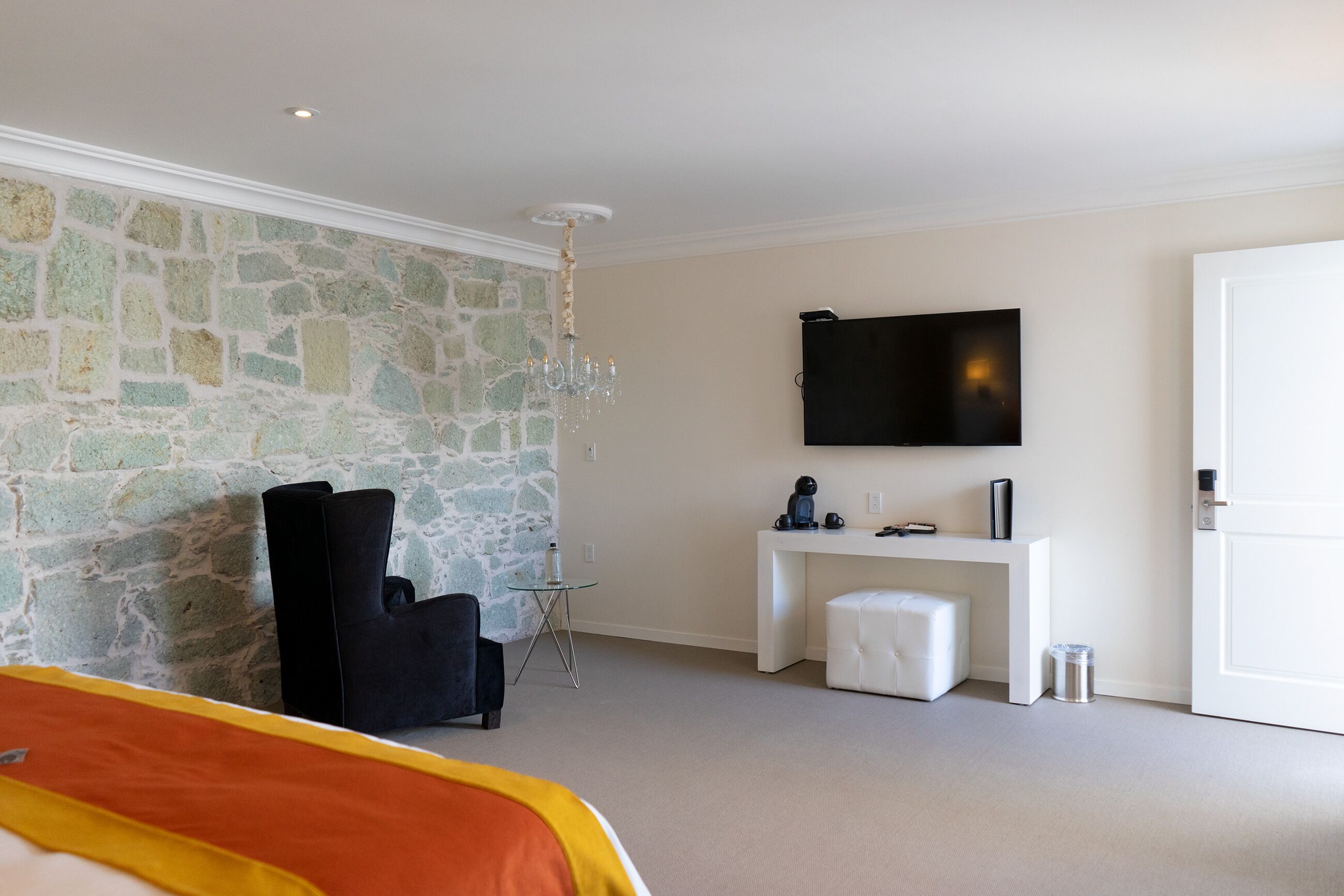 A hotel room with a stone wall, a bed with a yellow and orange blanket, a black chair, a small white table, a white bench, a TV mounted on the wall, and a door.