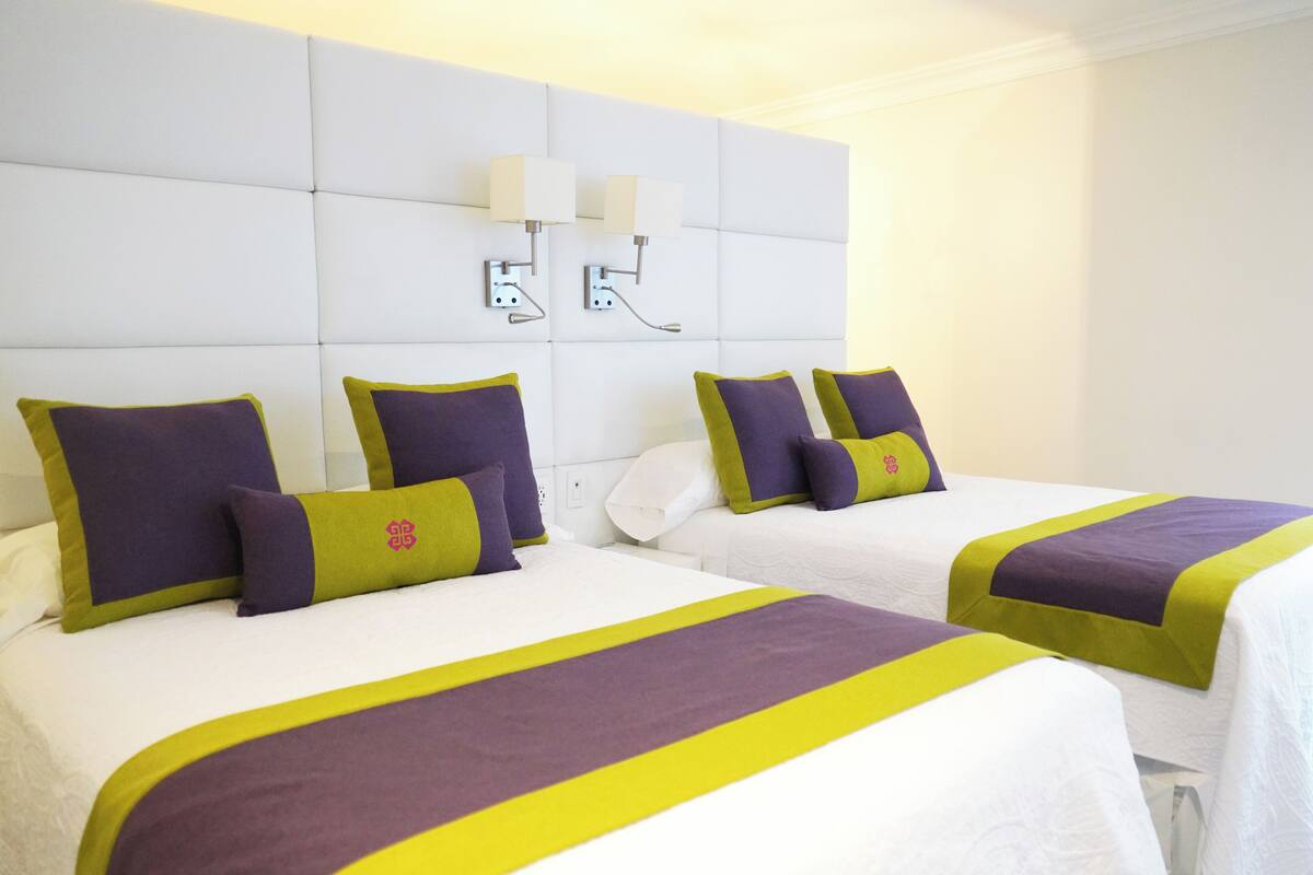 A hotel room with two beds, each with a white headboard, purple and green bedding, and a wall-mounted light fixture.