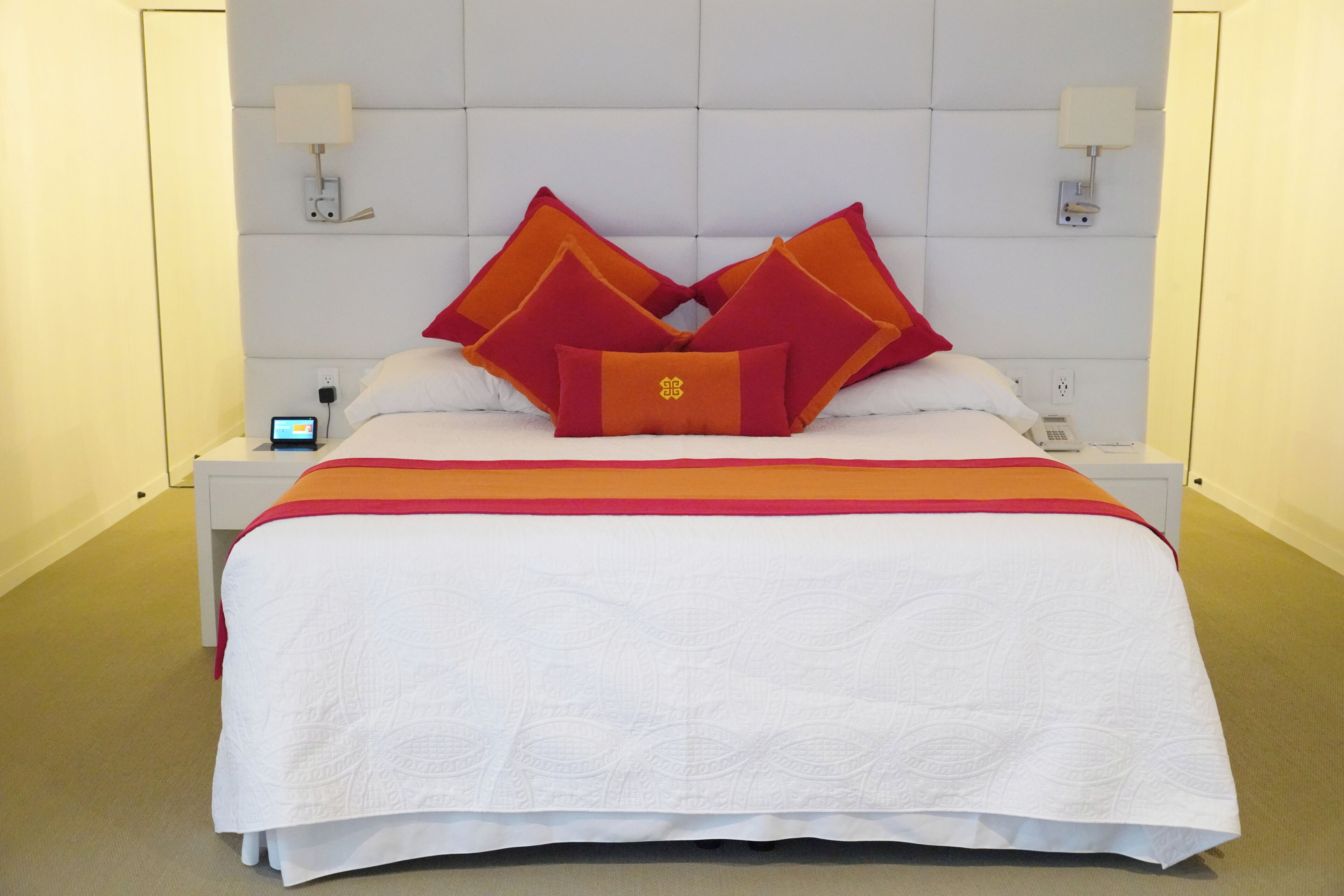 Une chambre d’hôtel avec un lit, du linge de lit blanc, une couverture à rayures rouges et oranges, et deux lampes de chevet.