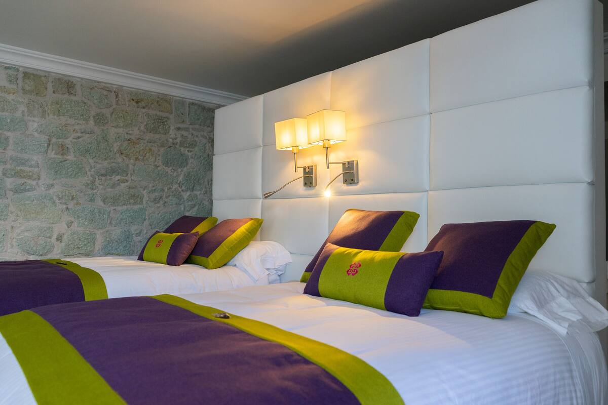 A hotel room with two beds, each with a white mattress and green and purple bedding. There is a stone wall on one side and a white padded headboard with a wall-mounted light fixture.