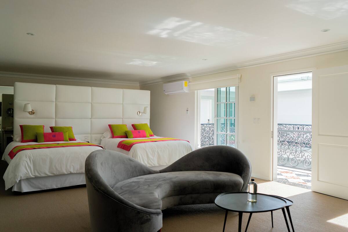 A hotel room with two beds, a grey armchair, a small table, and a balcony with a railing.
