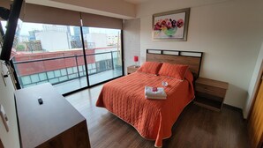 Habitación de hotel con una cama grande, una mesita de noche, un televisor y una ventana con vistas a la ciudad.