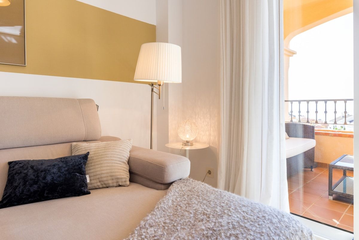 A hotel room with a sofa, a lamp, and a view of the outdoor balcony.