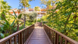 A wooden bridge leading to a building with balconies, surrounded by lush greenery.