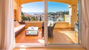 A balcony with a sofa, a table, and a chair, overlooking a scenic view of a coastal town.