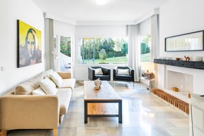A modern living room with a sofa, a coffee table, and two armchairs. There is a fireplace with a mantel and a large window providing a view of the garden.