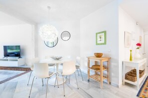 A modern living room with a flat-screen TV, a round dining table with chairs, a white sofa, a wooden side table with a bowl, and a wall-mounted mirror.