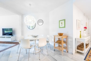 A modern living room with a white dining table, four white chairs, a TV, a chandelier, and a wooden side table with baskets.