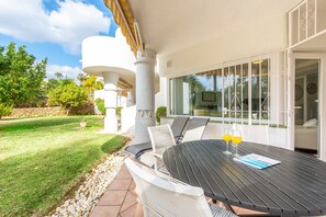 An outdoor patio with a round table and chairs, a small garden area, and a white building with a balcony.
