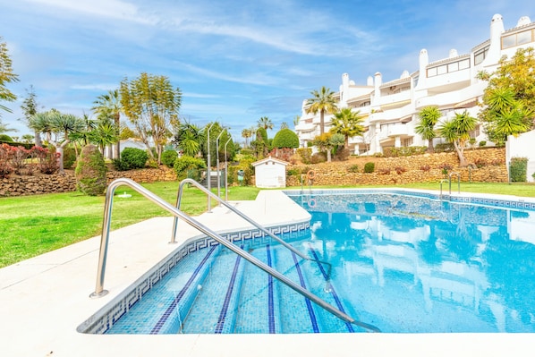 A swimming pool with a stone border, surrounded by a well-maintained garden and a white building with balconies.