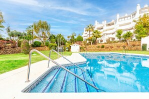 A swimming pool with a stone border, surrounded by a well-maintained garden and a white building with balconies.