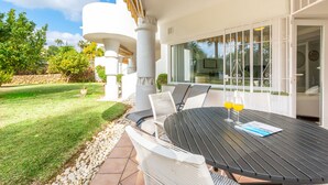 Un patio al aire libre con una mesa redonda, sillas de mimbre blanco y una puerta de vidrio que da a un área techada.