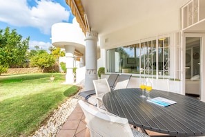 An outdoor patio with a round table, white wicker chairs, and a glass door leading to a covered area.