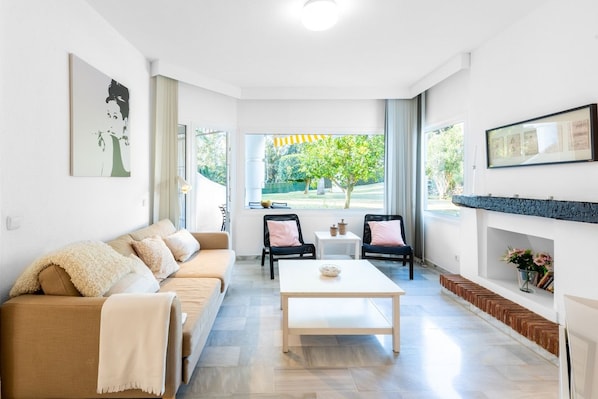 A modern living room with a sofa, coffee table, and chairs, a fireplace, and a view of a garden.