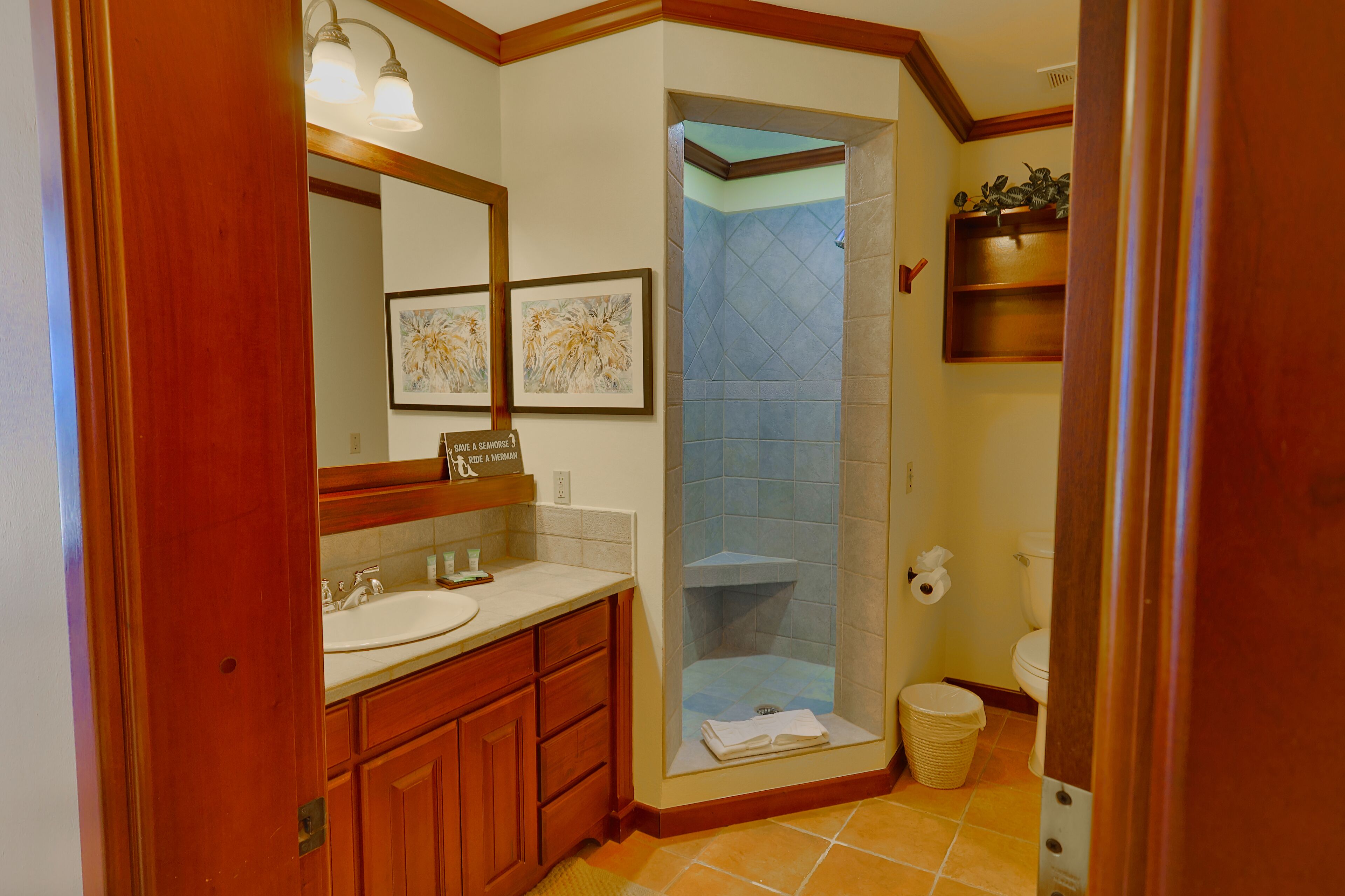 Een badkamer met een houten wastafelmeubel, een wastafel, een spiegel, een kunstwerk in een lijst, een douche met een blauwe betegelde wand, en een toilet.