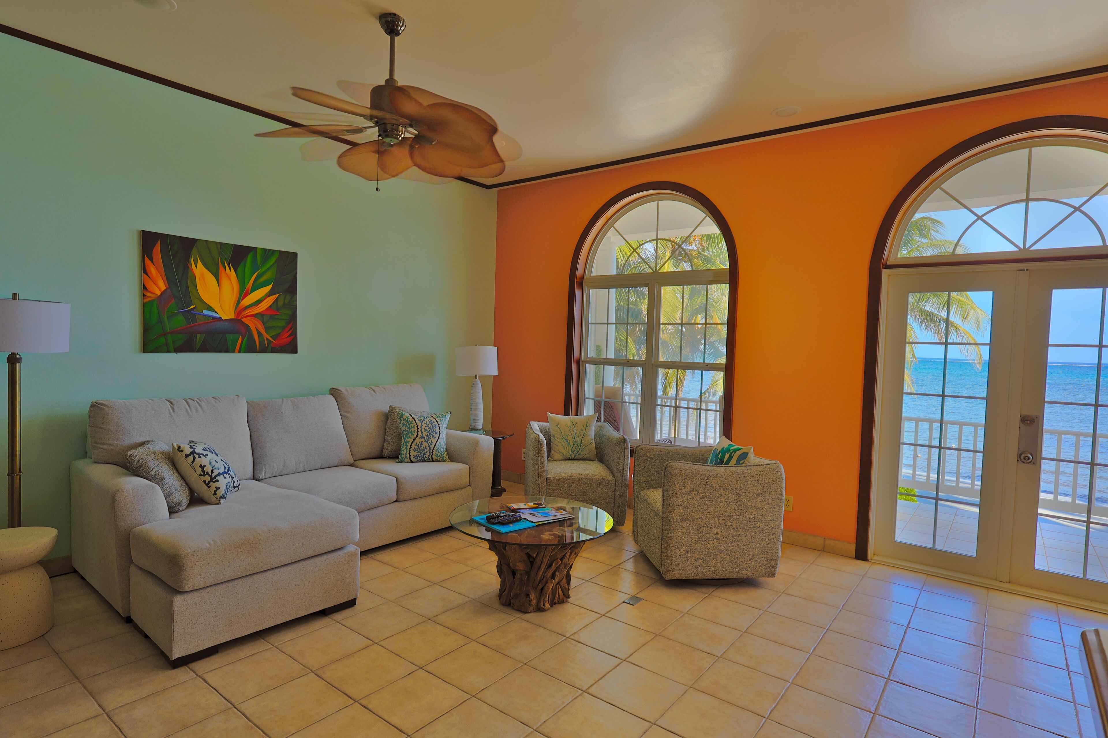 A living room with a beige sofa, a glass coffee table, and two armchairs. There is a ceiling fan, a painting of tropical flowers, and a view of the ocean through large windows.