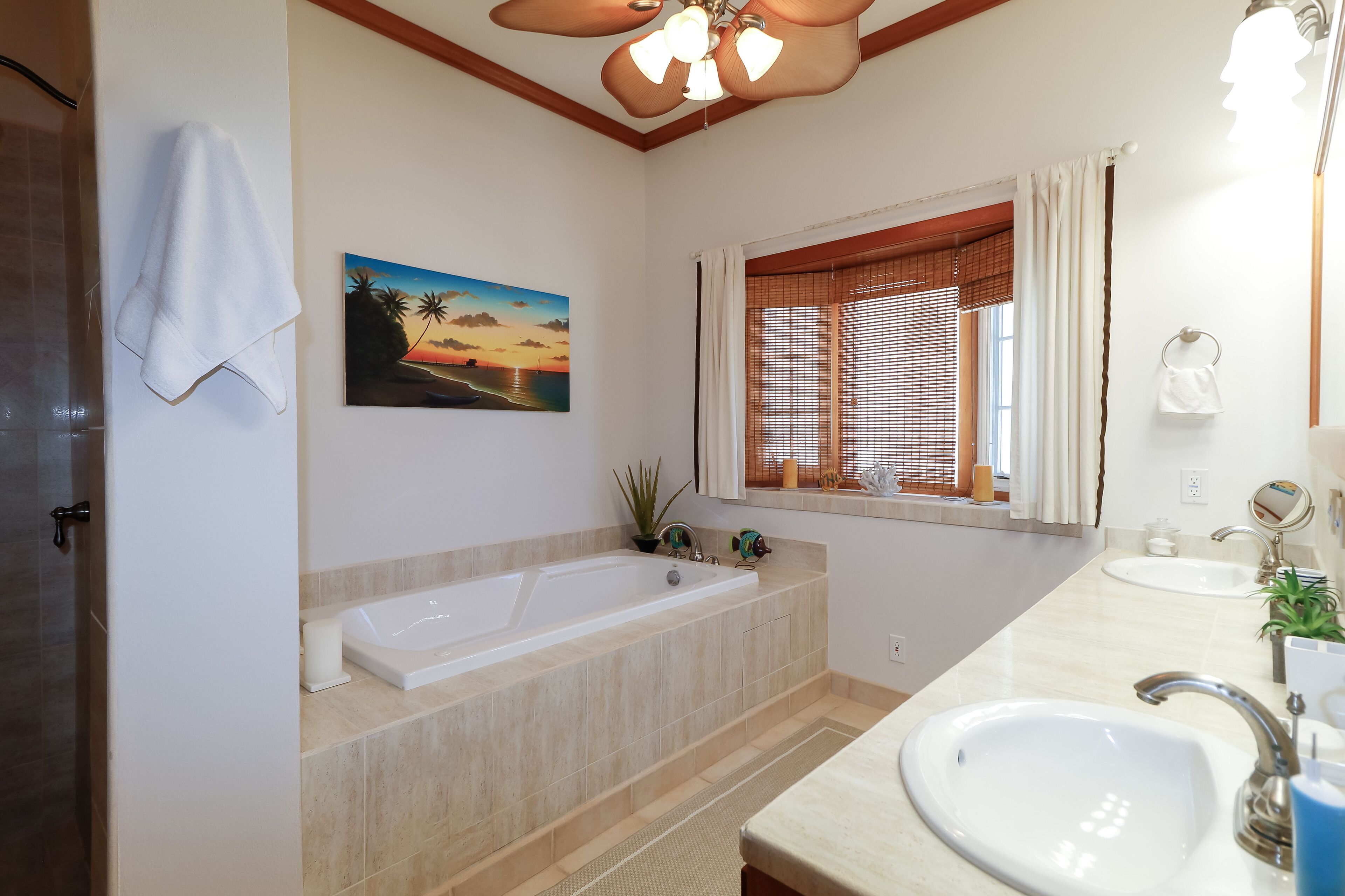 Ein Badezimmer mit Badewanne, Waschbecken, einem Fenster mit Jalousien, einem Handtuch an der Wand und einem Sonnenuntergangsbild.